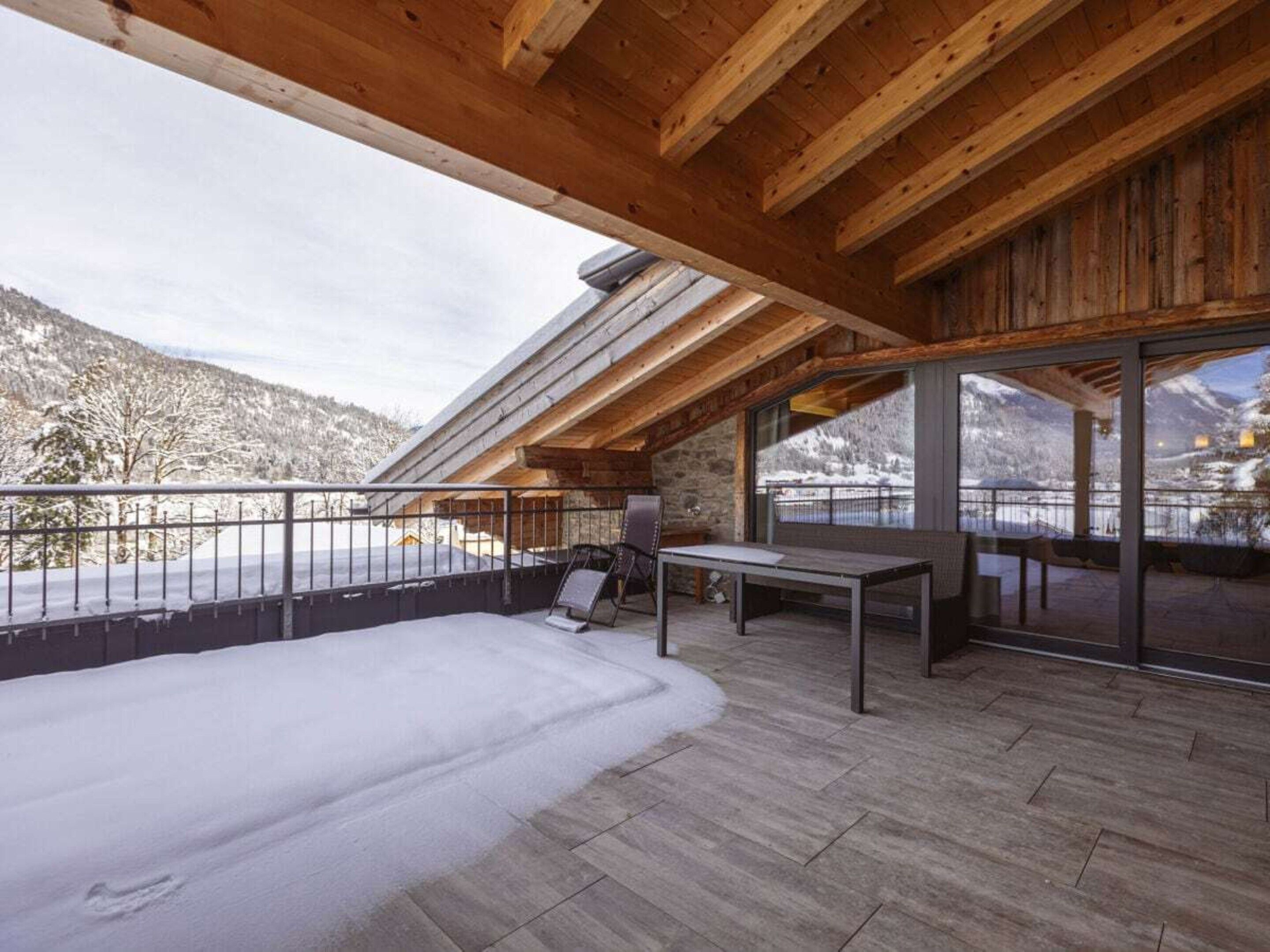 A snow-covered outdoor area with a wooden deck, a table, and chairs, overlooking a mountainous landscape.