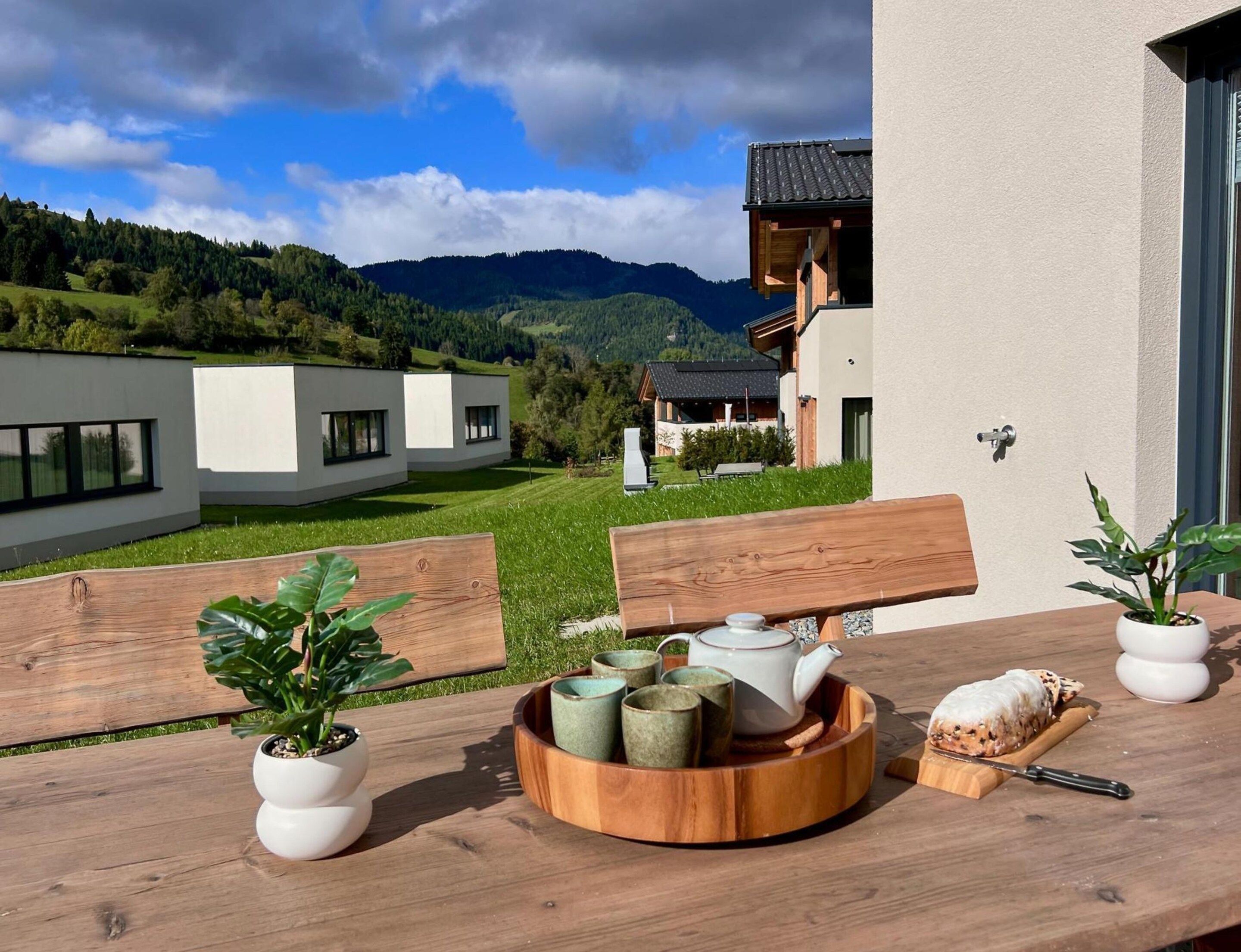 Una mesa de madera con un hervidor, tazas y una tabla de cortar, ubicada en un balcón con vistas a un edificio moderno y un paisaje montañoso.