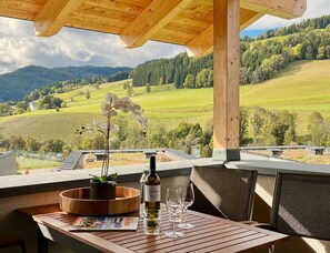 A wooden table with a bottle of wine, glasses, and a magazine, overlooking a scenic landscape with hills and trees. - Chalet Right Next to the ski Area (Sankt Lambrecht)