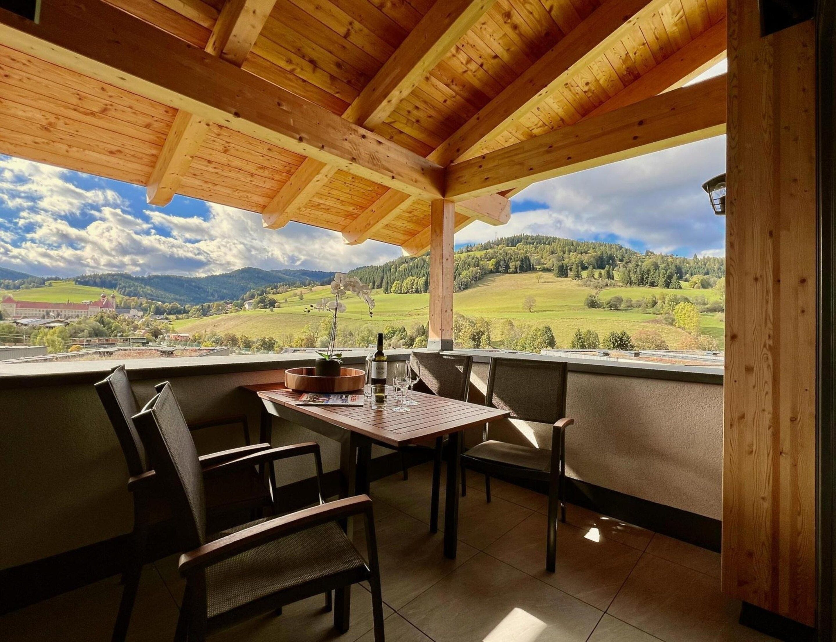 Ein Holzbalkon mit Tisch und Stühlen, der auf eine reizvolle Landschaft mit Hügeln und einem Gebäude in der Ferne blickt.