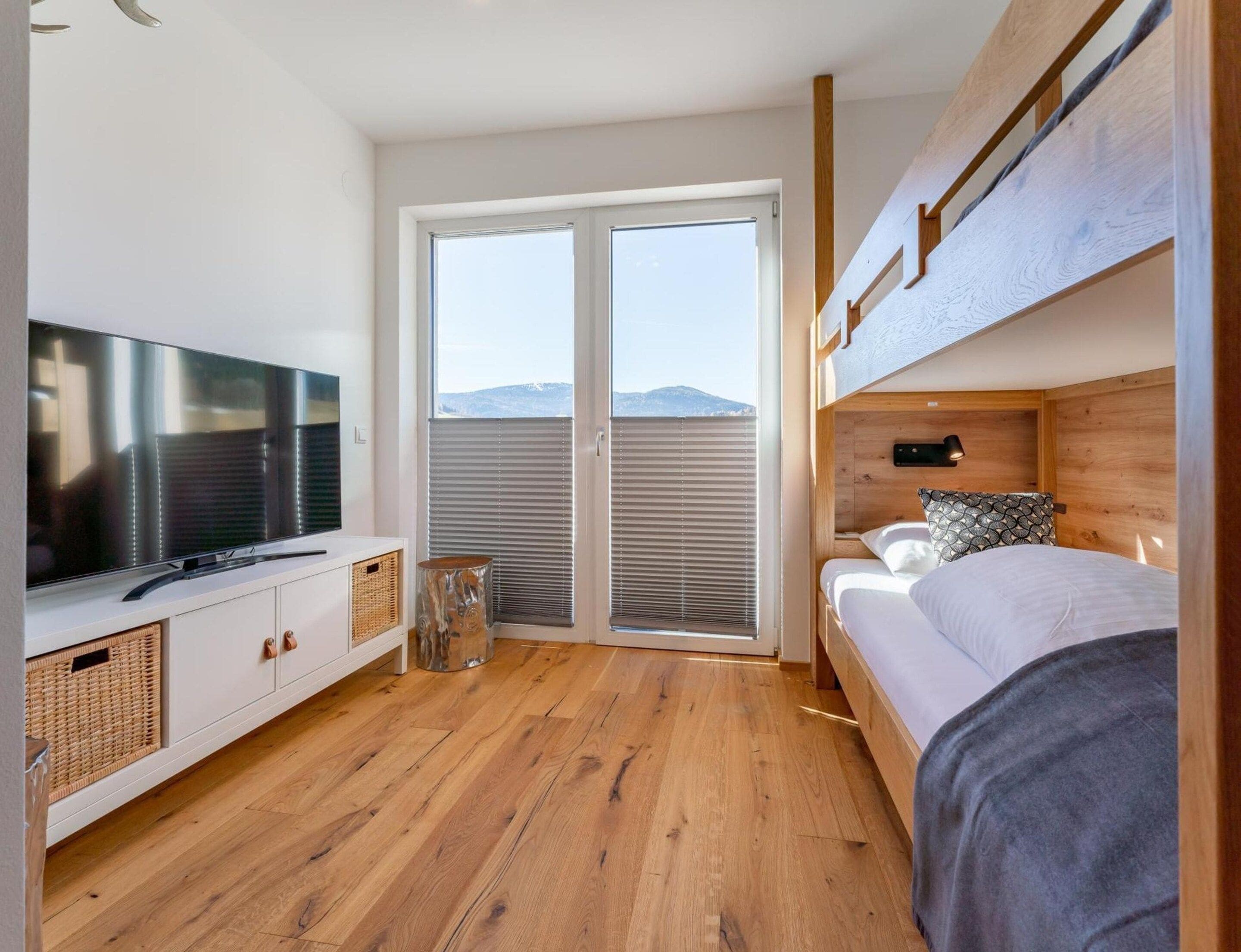 Un dormitorio moderno con televisión de pantalla plana, piso de madera, litera y puerta corrediza de vidrio que da a un balcón.