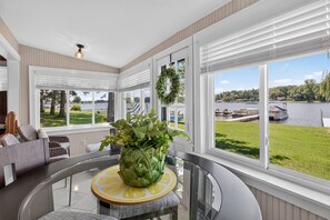 A modern interior with a glass table, a potted plant, and a view of a lake with boats. - Lakefront Cottage w/ Sunset Views and Private Dock (Brooklyn)