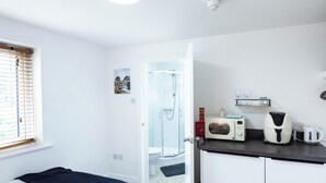 Quarto de hotel compacto com cama, chuveiro, cozinha compacta equipada com micro-ondas e chaleira, e janela com persianas.