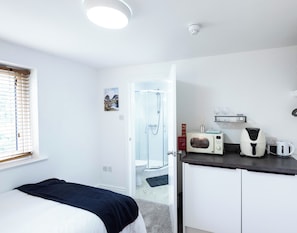 A compact hotel room with a bed, a shower, a kitchenette with a microwave and kettle, and a window with blinds. - The Stanley Arms Hotel by Guestz (Seascale)