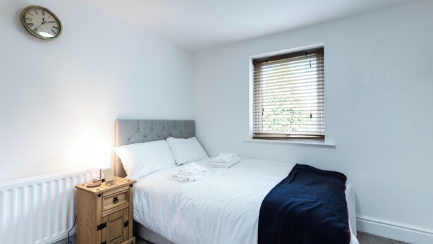 Quarto com cama, mesa de cabeceira, janela com persianas e relógio na parede.