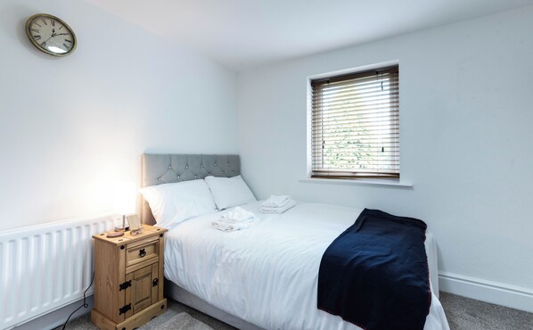 A bedroom with a bed, a bedside table, a nightstand, a window with blinds, and a wall clock. - The Stanley Arms Hotel by Guestz (Seascale)