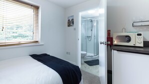 Quarto com cama, janela com persianas, área de chuveiro e micro-ondas em bancada.