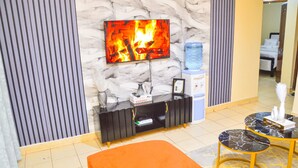 Vestíbulo de hotel con un televisor de pantalla plana mostrando un fuego, un dispensador de agua, una consola negra con un cuadro enmarcado y una mesa redonda con un libro de Louis Vuitton.