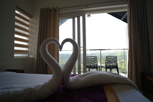 A hotel room with a bed, a heart-shaped towel arrangement, a window with curtains, and a view of a balcony with chairs.
