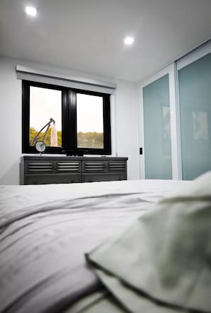 Ein modernes Schlafzimmer mit Bett, Kommode, Fenster mit Blick und einer Schiebetür.
