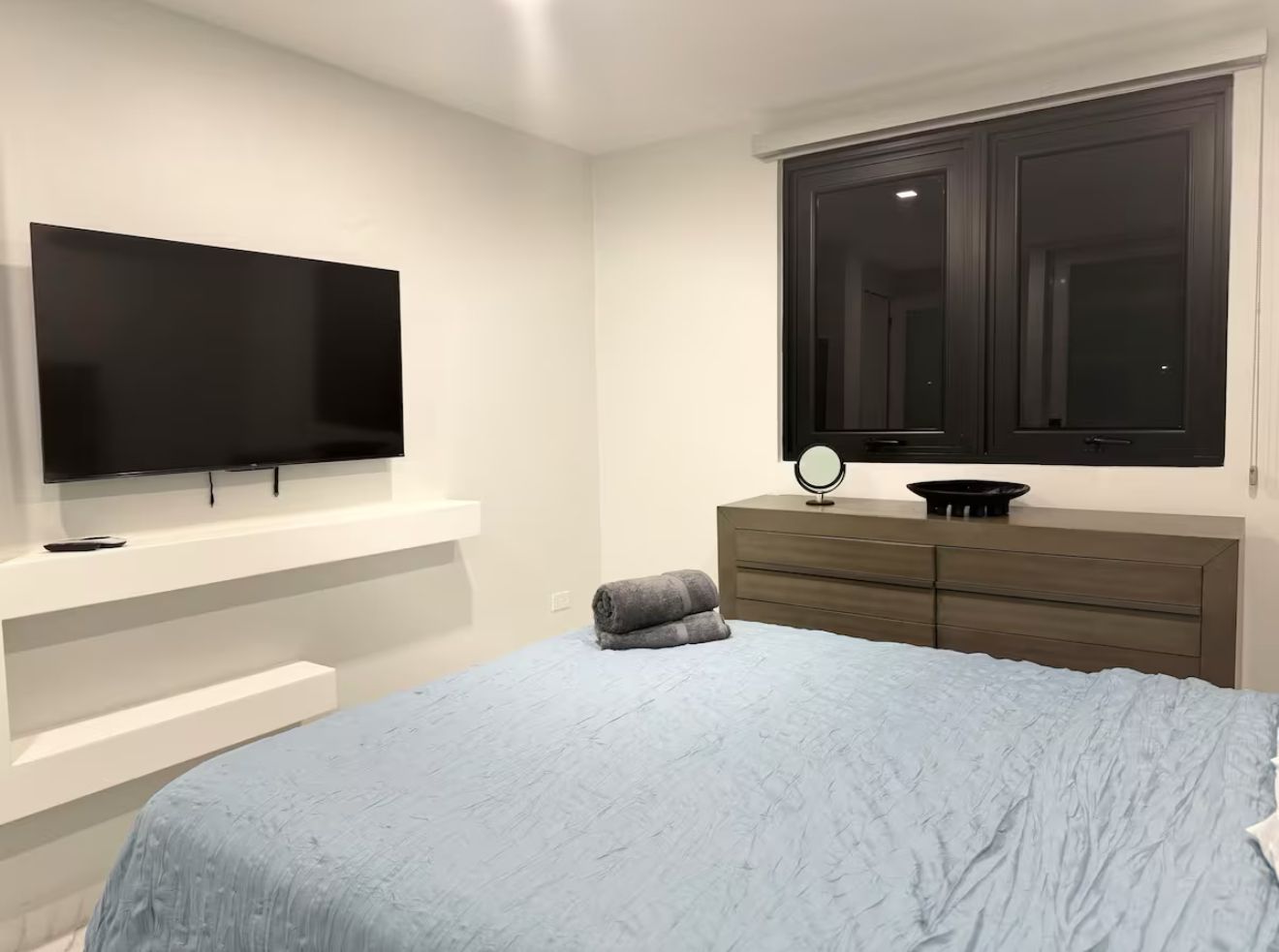 A bedroom with a bed, a television mounted on the wall, a dresser, and a window with dark curtains.