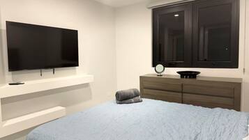 A bedroom with a bed, a television mounted on the wall, a dresser, and a window with dark curtains.