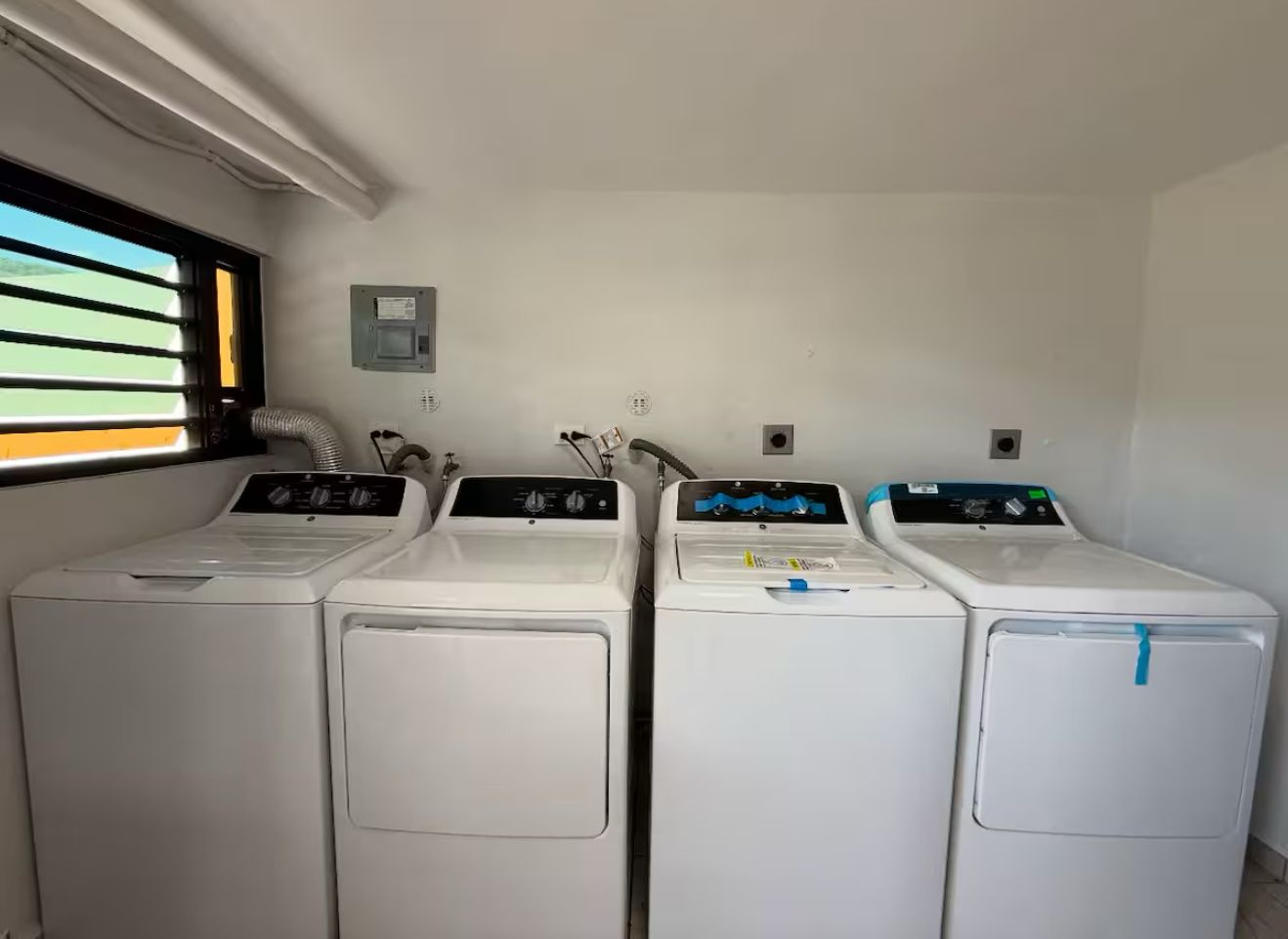 A laundry room with four white front-loading washing machines, a window with blinds, and a small electrical panel on the wall.