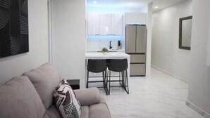 Um interior moderno com sofá, área de jantar para duas pessoas, cozinha com armários brancos e geladeira.