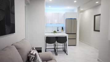 A modern interior with a sofa, a dining area with two chairs, a kitchen with white cabinets, and a refrigerator.