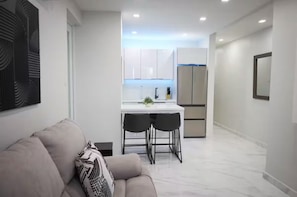 Ein modernes Interieur mit einer Couch, einem Essbereich mit zwei Stühlen, einer Küche mit weißen Schränken und einem Kühlschrank.