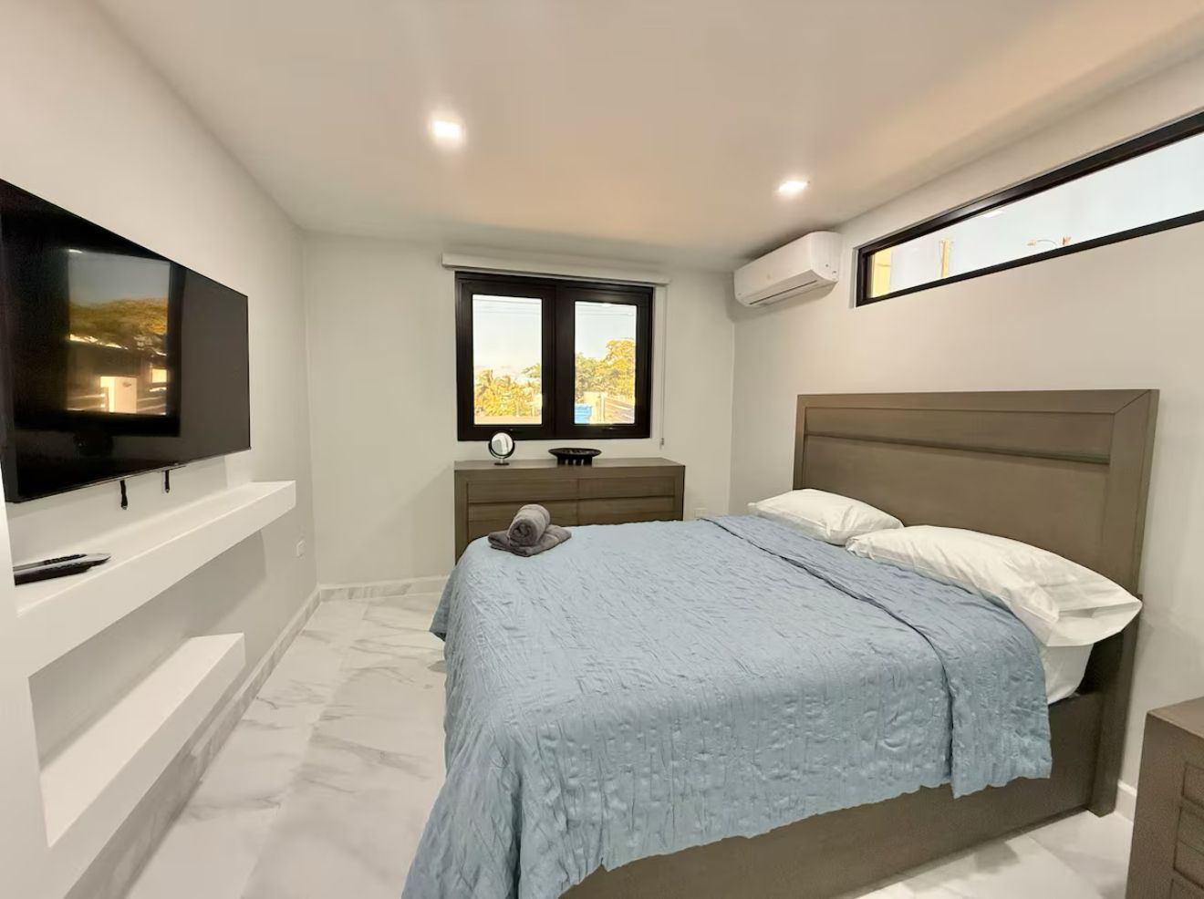 Un dormitorio con cama, televisión sobre un mueble, ventana y mesita de noche.