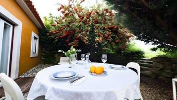 An outdoor dining area with a table set for two, surrounded by a stone wall and a bush with red berries.