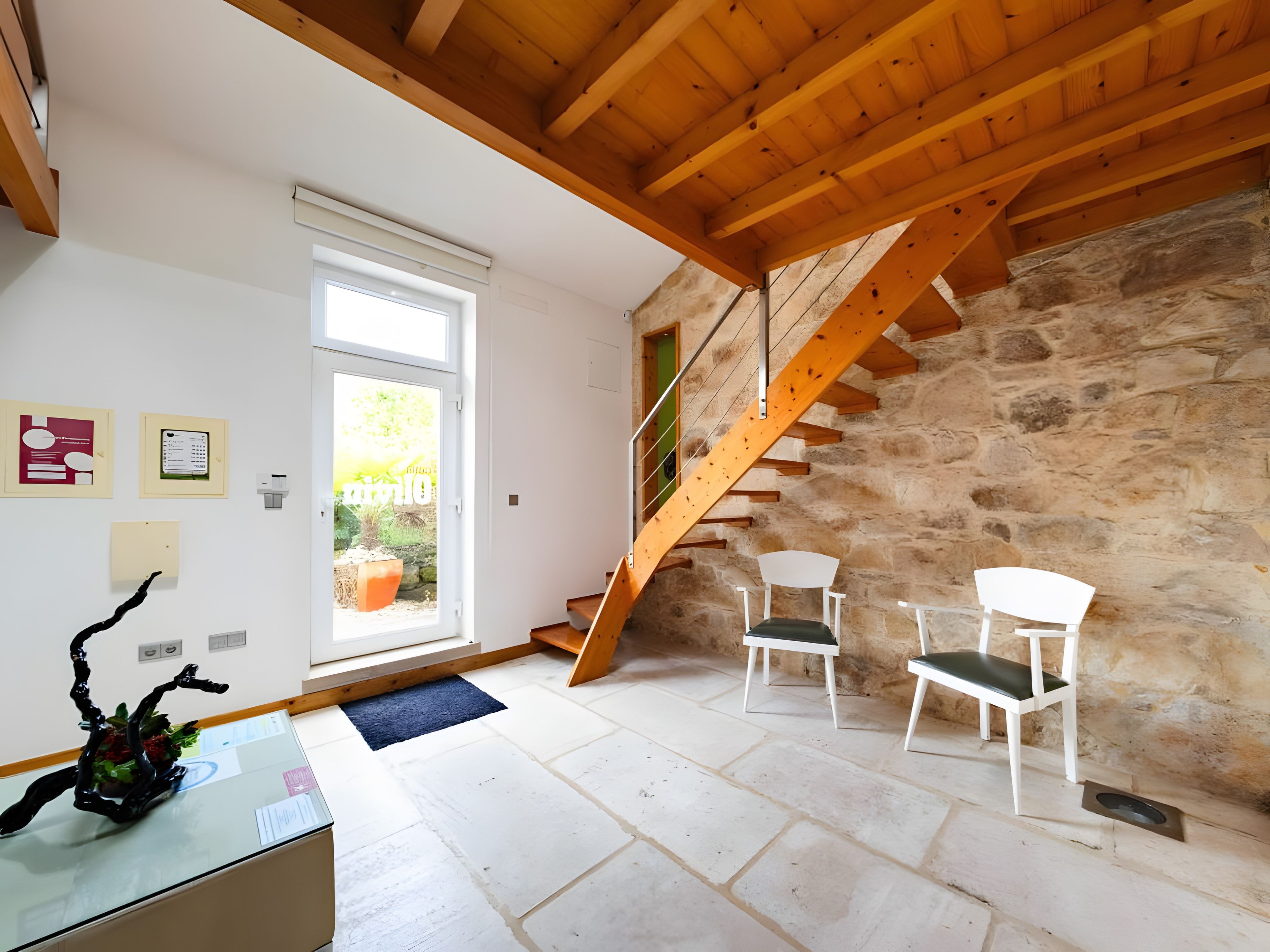 Une pièce avec un mur en pierre, un escalier en bois et deux chaises blanches.