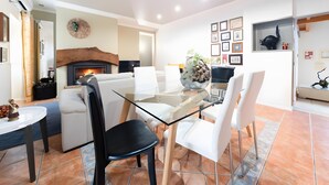 A modern dining area with a glass table, white chairs, a fireplace, and a wall decorated with framed pictures. - Country House 'D’Olívia Lovely Homes -Casa Olívia' with Shared Garden, Wi-Fi and Air Conditioning (Cela)