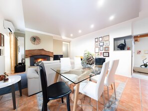 Een moderne eetruimte met een glazen tafel, witte stoelen, een open haard en een muur versierd met ingelijste foto's.