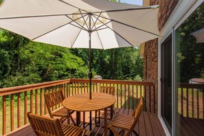 Eine Holzterrasse mit Tisch, Stühlen und Sonnenschirm, sowie eine Glastür, die zu einem Garten führt.