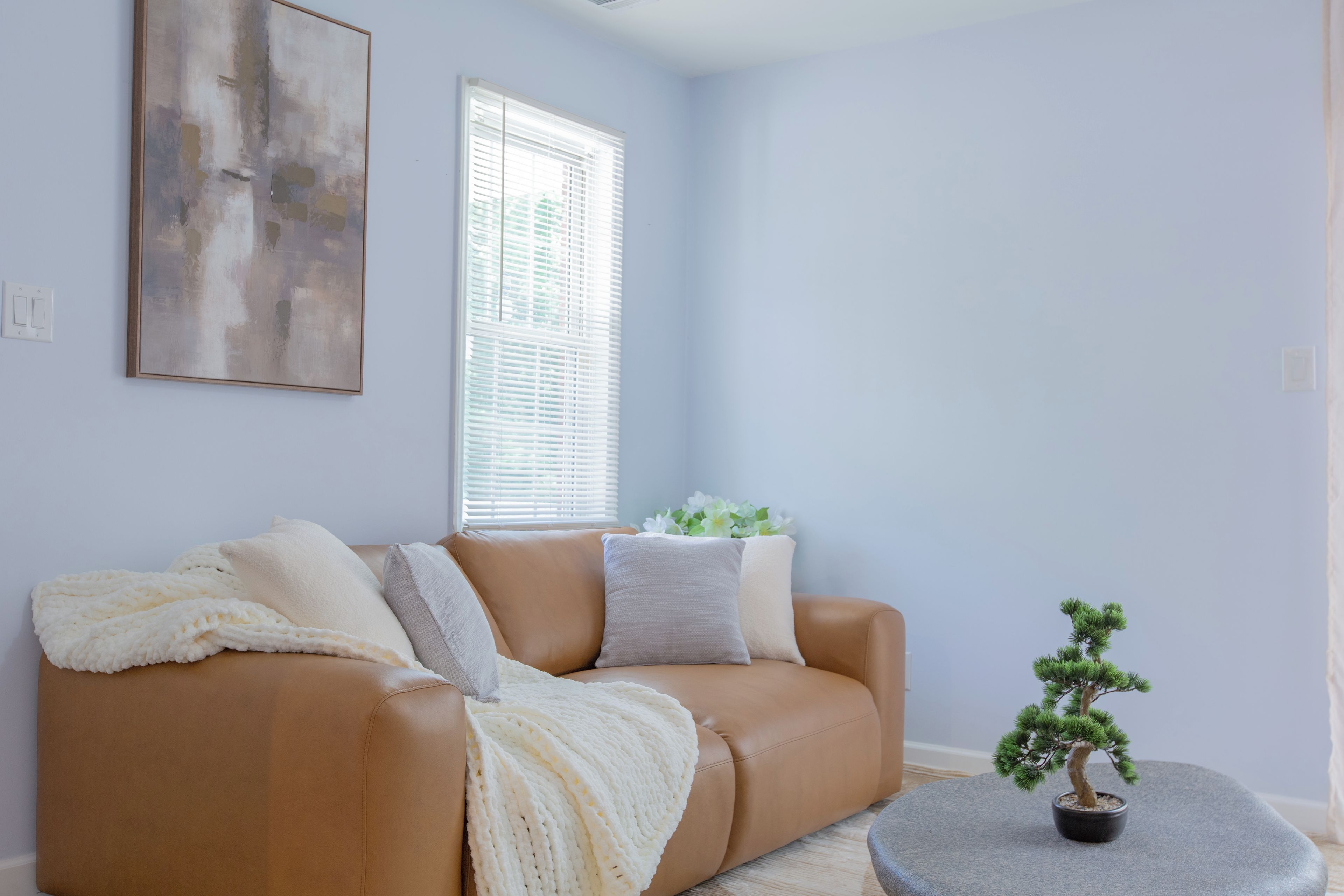 Sala de estar com um sofá de couro marrom, um cobertor branco de tricô, uma pequena árvore bonsai em vaso e um grande quadro abstrato na parede.