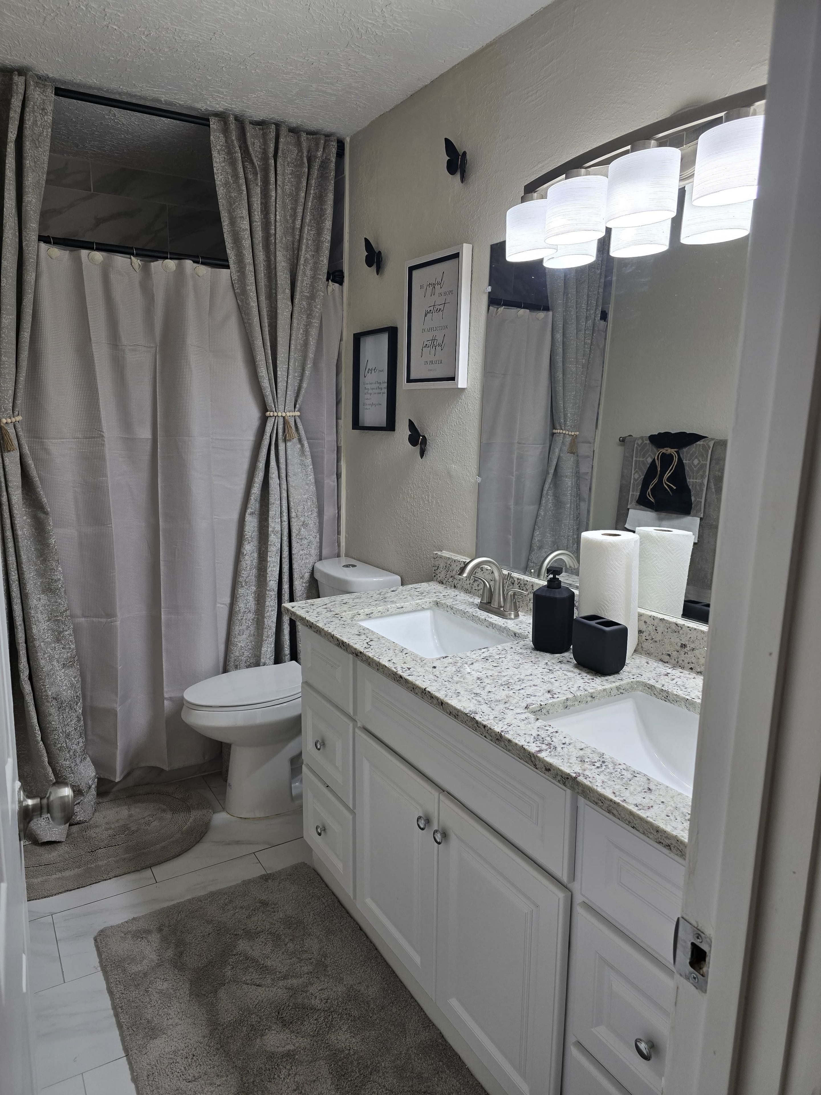 Une salle de bain avec des toilettes blanches, un lavabo doté d’un plan de travail en granit, un miroir et un rideau de douche.