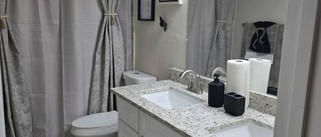 Une salle de bain avec des toilettes blanches, un lavabo doté d’un plan de travail en granit, un miroir et un rideau de douche.