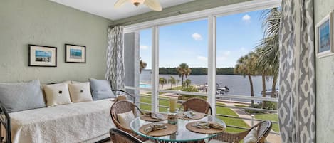 A room with a bed, a dining table with chairs, a ceiling fan, and a view of a lake with palm trees.