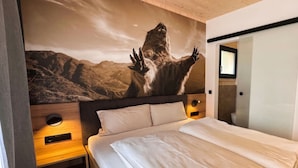 Un dormitorio con una cama grande, un cabecero con un mural de paisaje montañoso, una mesita de noche con una lámpara y una puerta que da a un baño.