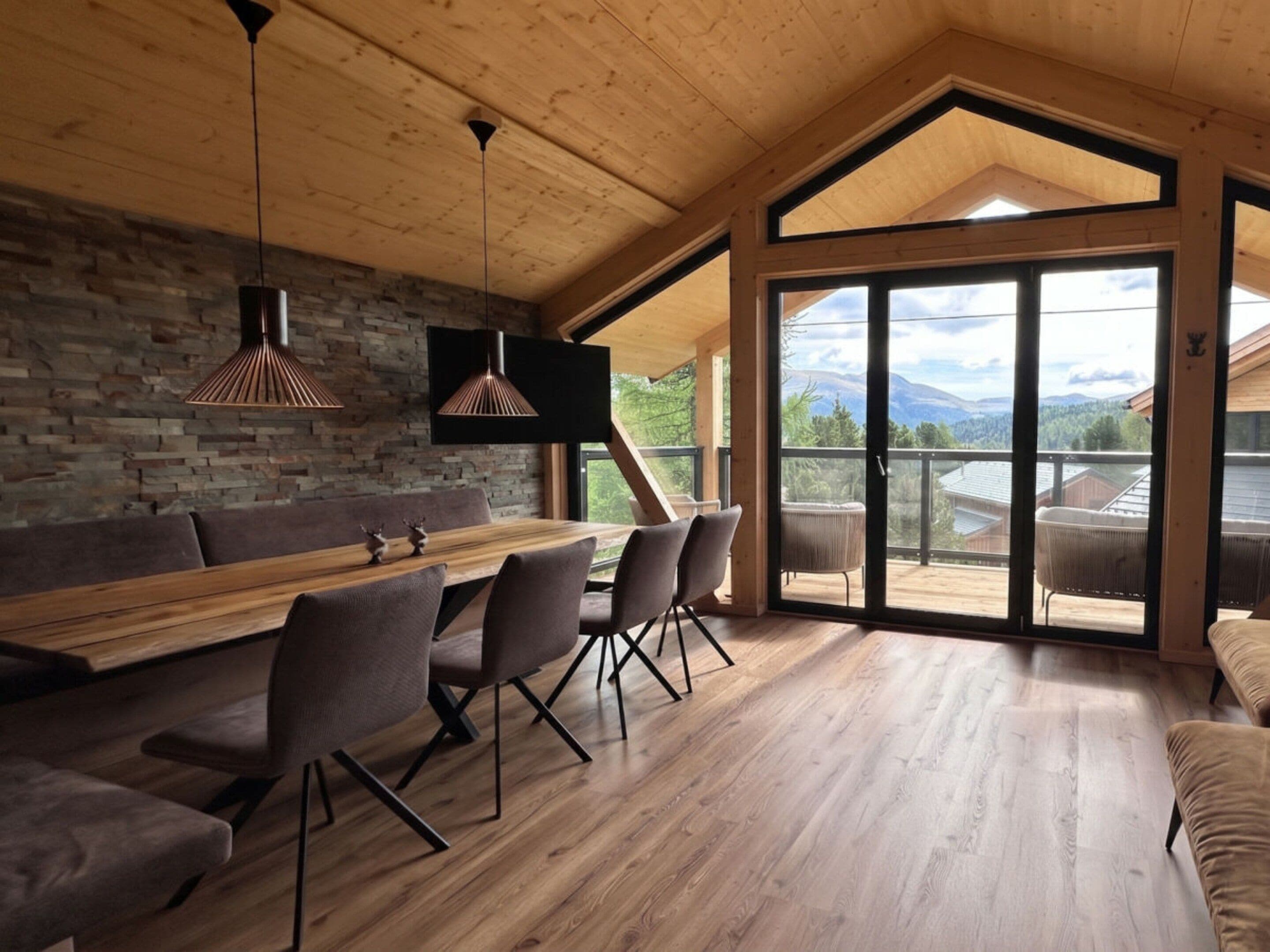 Un'area pranzo moderna con tavolo e sedie in legno, una parete in pietra e un'ampia porta scorrevole in vetro che dà su un balcone con vista sulle montagne.