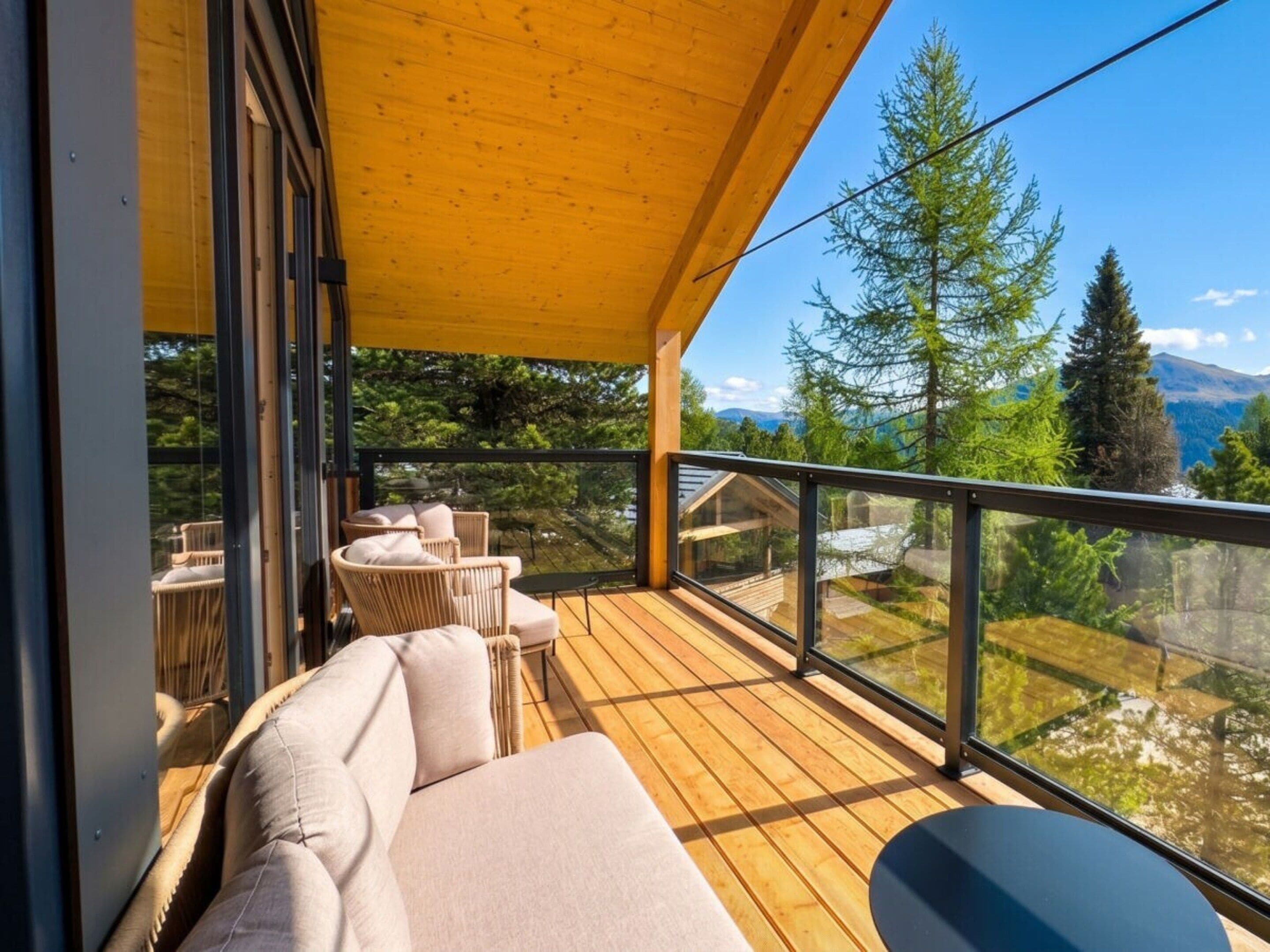Une terrasse en bois, dotée de mobilier d’extérieur, d’une balustrade en verre et offrant une vue sur les arbres et les montagnes.