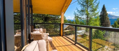 A wooden deck with outdoor furniture, a glass balustrade, and a view of trees and mountains.