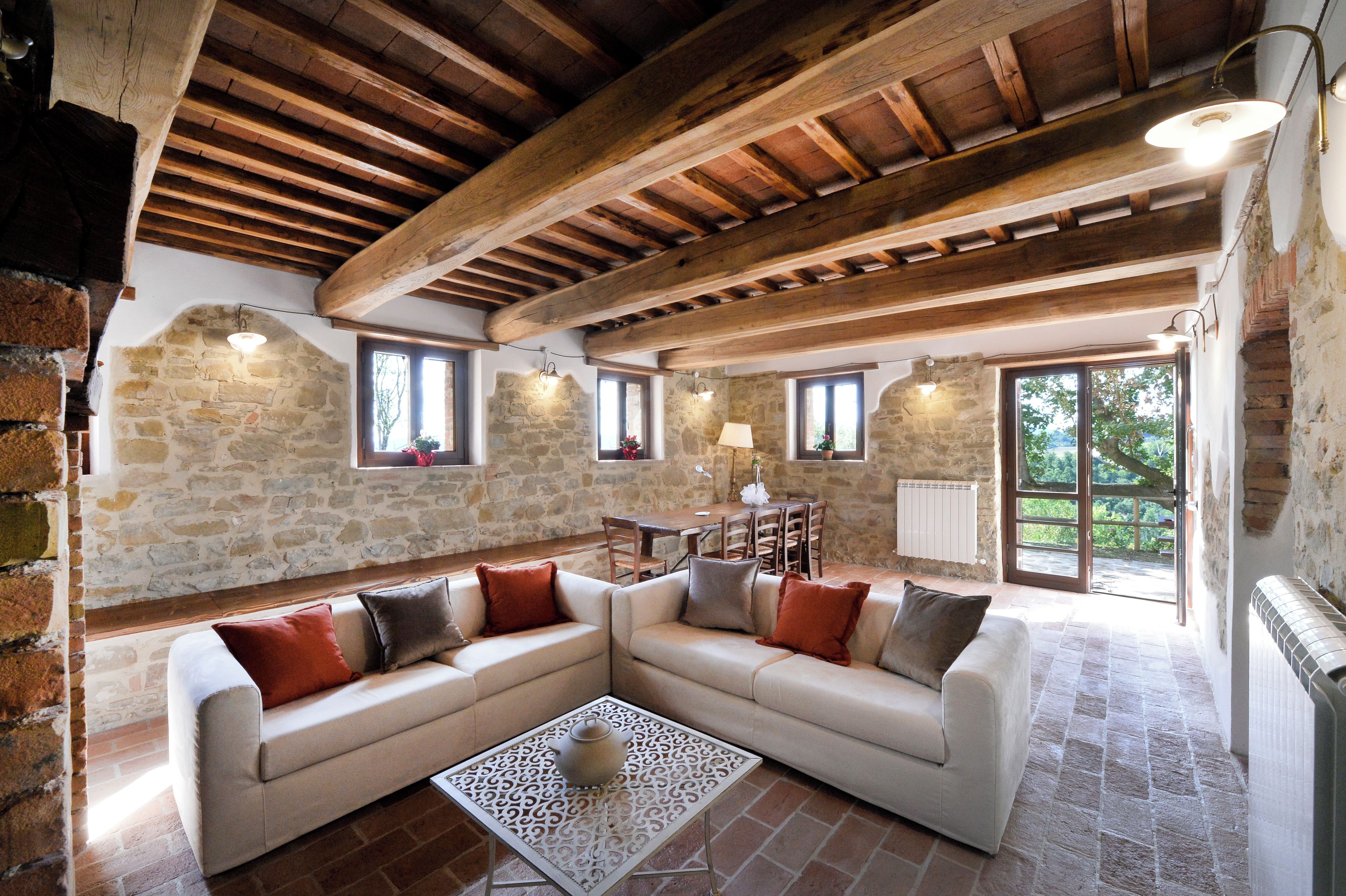 A rustic interior with exposed wooden beams, stone walls, a sectional sofa, and a dining area.