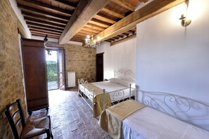A rustic room with a stone wall, wooden ceiling beams, a bed with a metal frame, a chair, and a window with a view of greenery. - Charming Italian Farmhouse with Sunset views, Ensuites & Private Infinity Pool (Umbria)