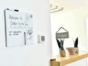 A whiteboard with a welcome message and WiFi details, a thermostat, and a shelf with plants and a sign. - Cedar By The Sea - steps to the beach/Flagler Ave (New Smyrna Beach)