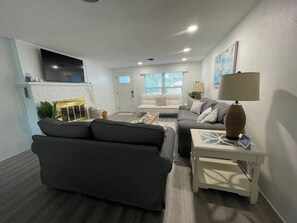 A living room with a sofa, a coffee table, a lamp, and a TV mounted on the wall. - Cedar By The Sea - steps to the beach/Flagler Ave (New Smyrna Beach)