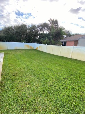 A backyard with a white fence, a grassy lawn, and trees in the background. - Sandy Toes on Cedar - steps from the beach (New Smyrna Beach)