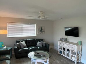 En stue med en sort lædersofa, et sofabord i glas, et hvidt konsolbord med skuffer, et fladskærms-tv på væggen og en loftsvifte.