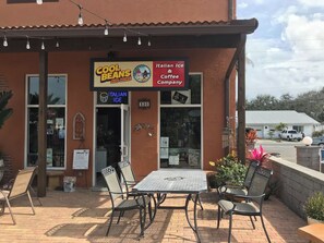 Un área de estar al aire libre con mesas y sillas, un letrero de Italian Ice & Coffee Company y una fachada con un letrero de Cool Beans y más.