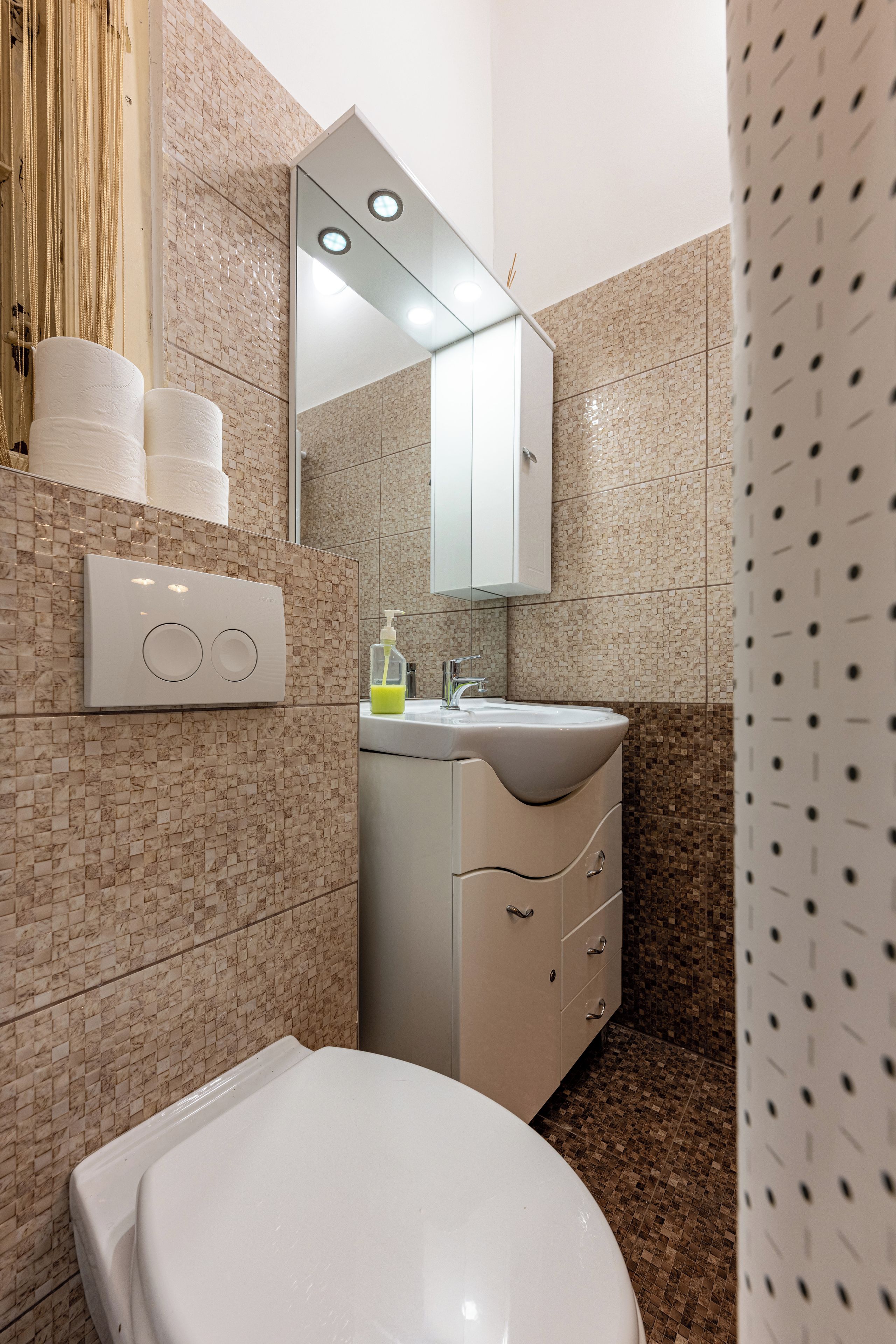 A bathroom with a toilet, a sink, and a mirror. There are towels and a soap dispenser on the wall.
