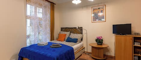 Un dormitorio con cama, televisión, una mesita con una planta y un cuadro en la pared.