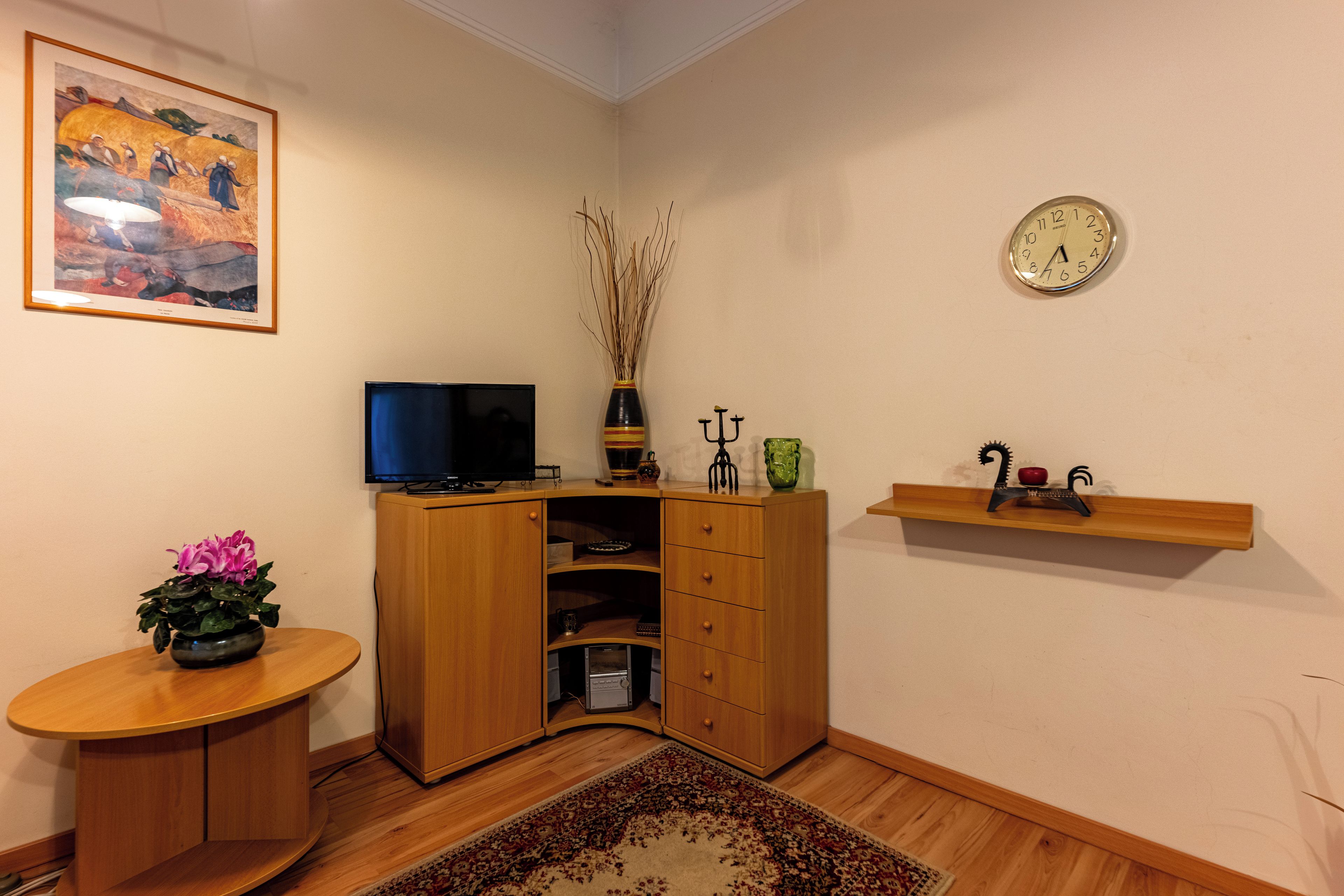 Un soggiorno con televisore a schermo piatto, mobiletti in legno, vaso decorativo, orologio e un tavolino con una pianta.