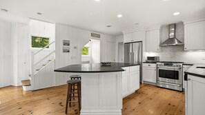 A modern kitchen with white cabinetry, a central island, stainless steel appliances, and a wooden staircase. - Updated Montauk retreat with breathtaking ocean views and effortless... (Montauk)