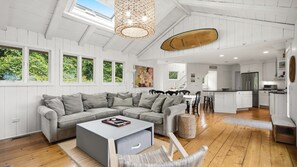 A modern living room with a grey sectional sofa, a coffee table, and a wicker chair. There is a kitchen area with white cabinetry and a wooden countertop. - Updated Montauk retreat with breathtaking ocean views and effortless... (Montauk)