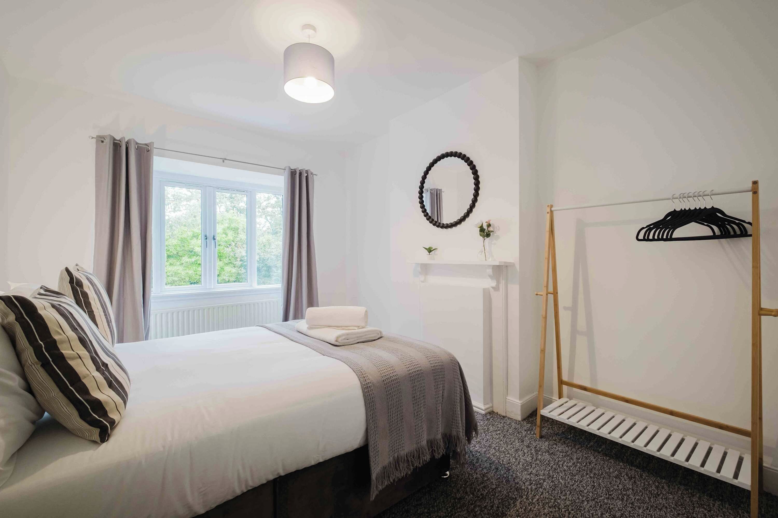 A hotel room with a bed, a window with curtains, a radiator, a wall-mounted mirror, a wooden hanger rack, and a radiator vent.