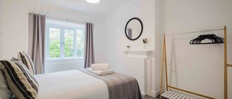 A hotel room with a bed, a window with curtains, a radiator, a wall-mounted mirror, a wooden hanger rack, and a radiator vent.