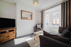 A modern living room with a flat-screen TV on a wooden cabinet, a sofa, a coffee table, and a large window with curtains. - 4 Bedroom House With 2 Bathrooms Near Town Centre (Northampton)