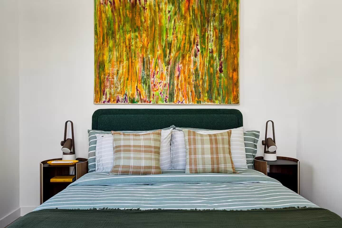Un lit recouvert d’une couvre-lit à rayures, deux tables de chevet avec des lampes et un grand tableau abstrait au-dessus du lit.